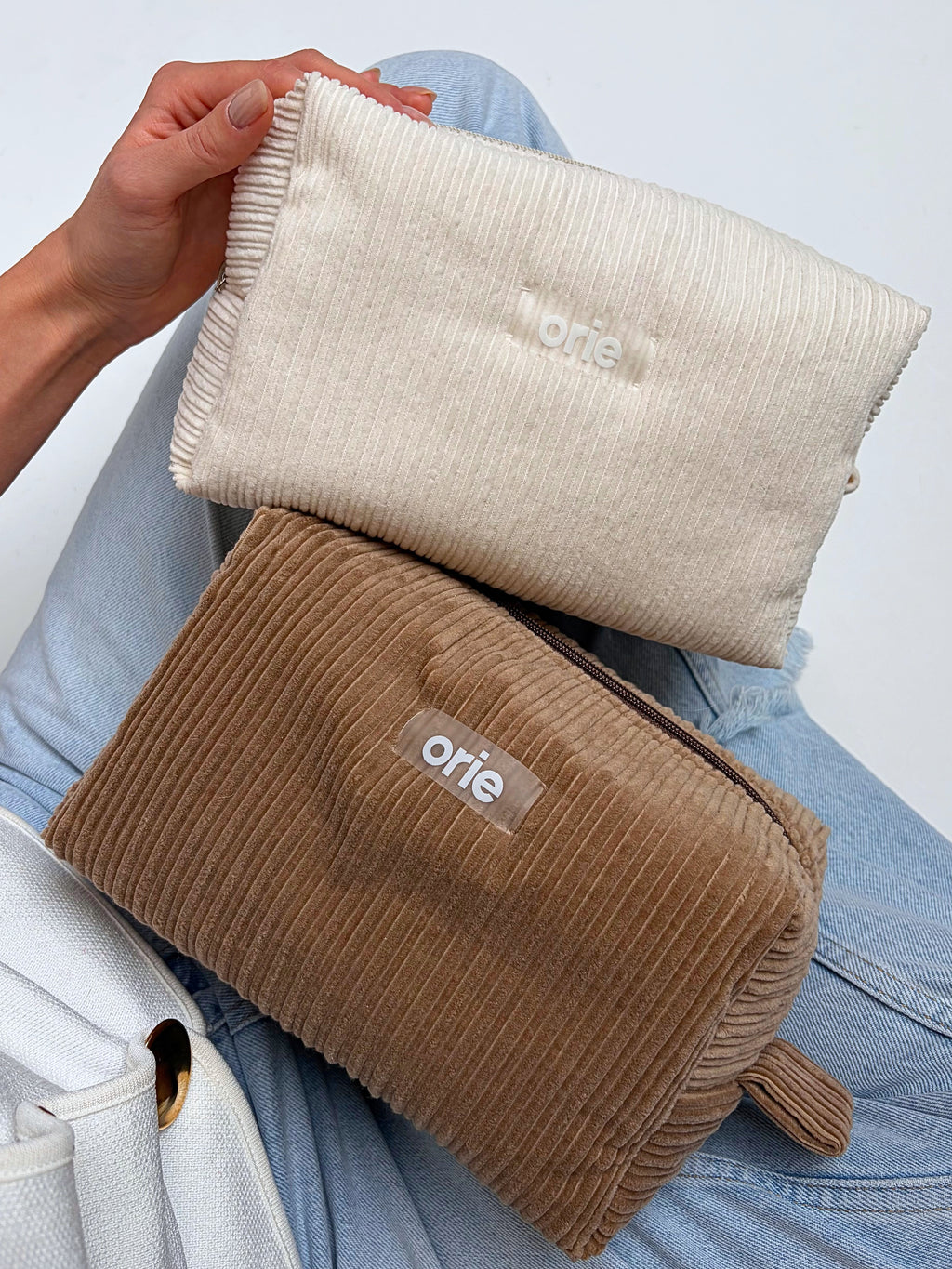 Two travel makeuopbags, one beige and one brown, held by a person wearing blue jeans.