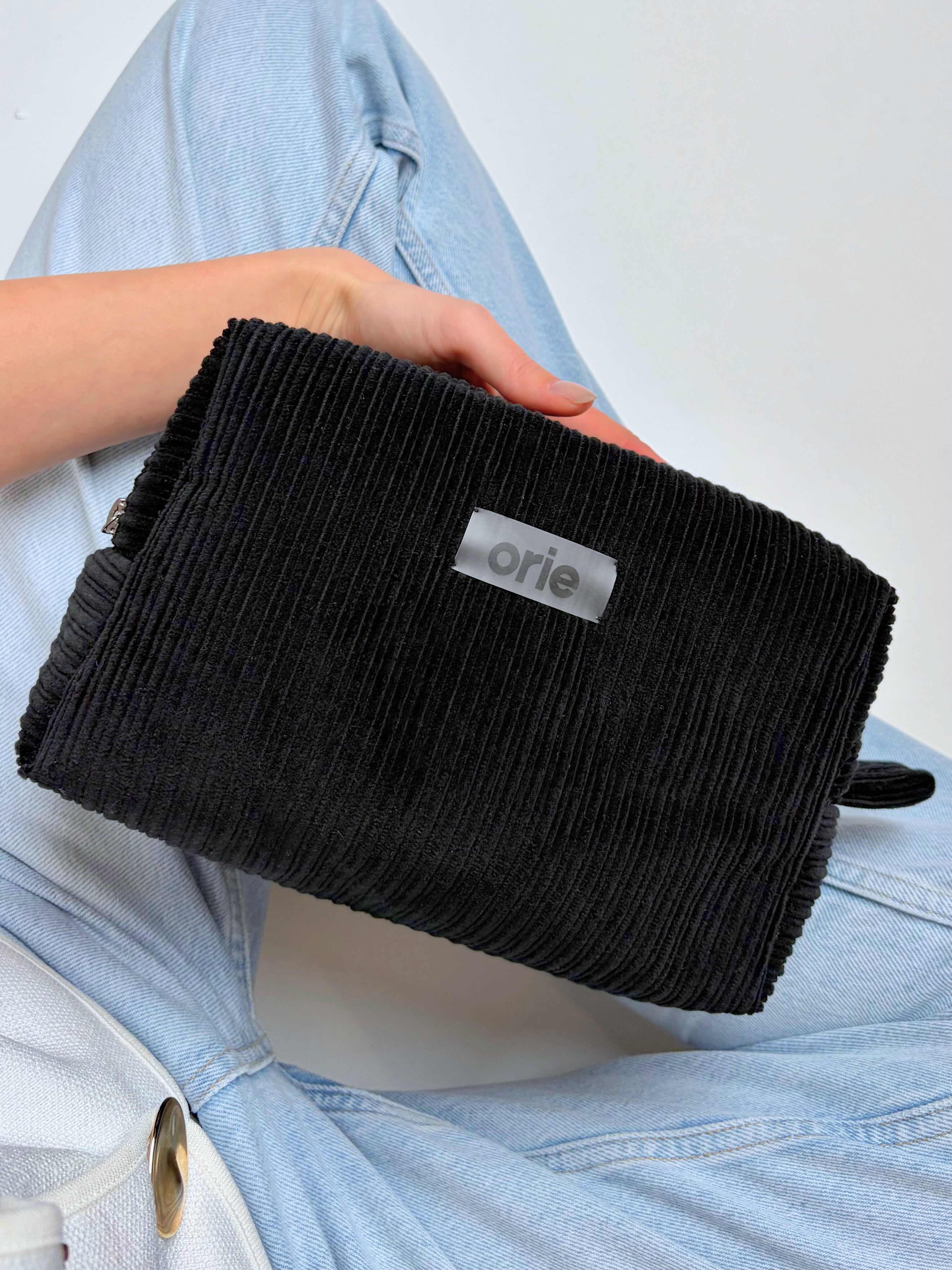 Black textured makeup bag held by a person wearing light blue jeans on a white background
