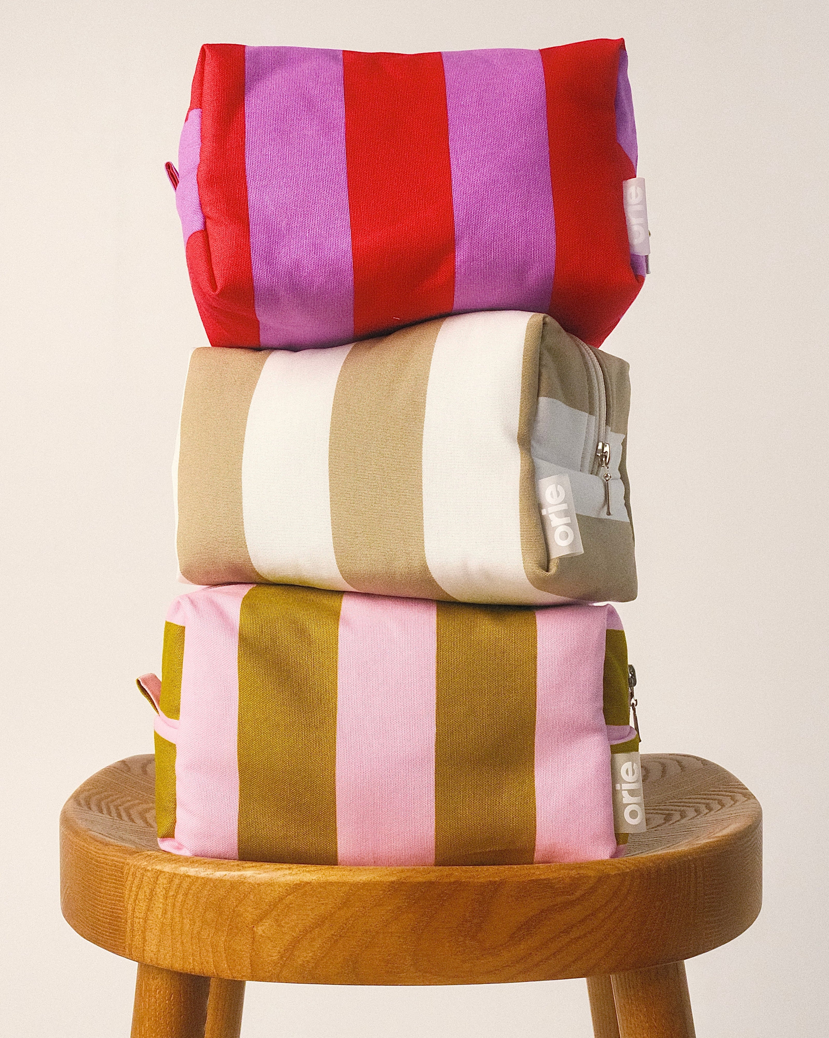 Stack of colorful striped bags on a wooden stool against a plain, background, orie kosmetinės/makeup bag