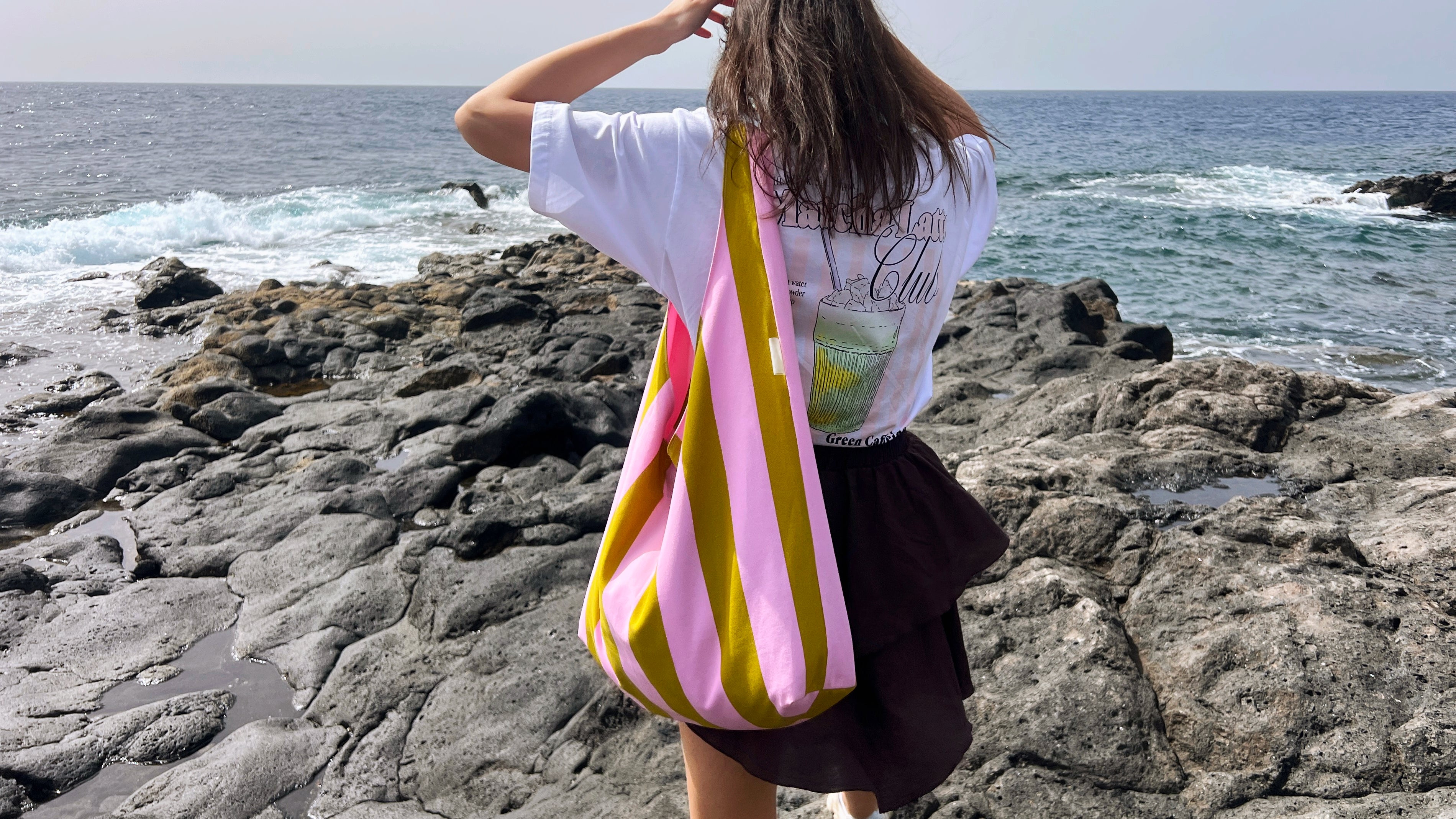Person holding a striped bag on a rocky beach with ocean in the background orie brand green and pink