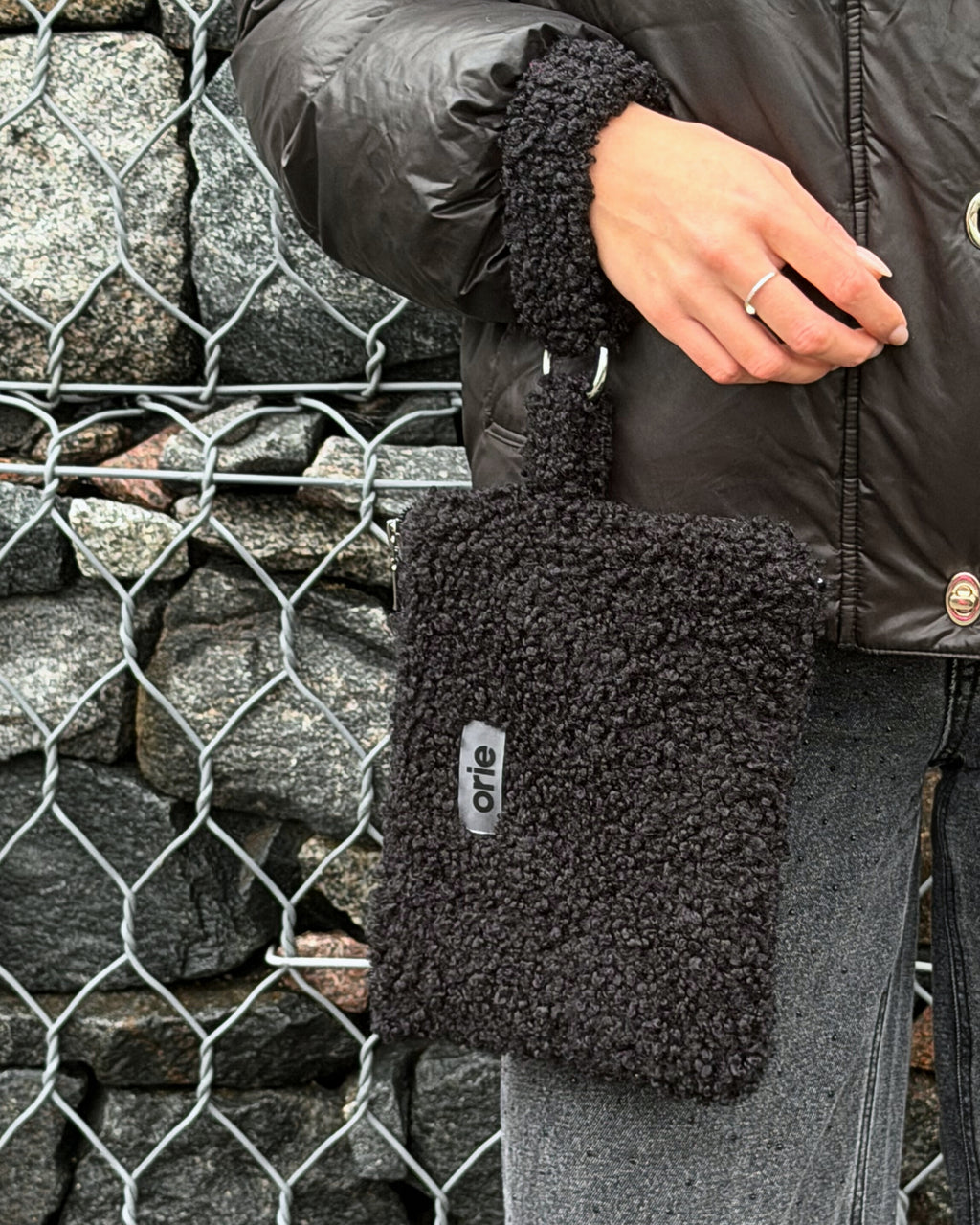 Person holding a black teddy bag with a visible brand logo against a stone wall.