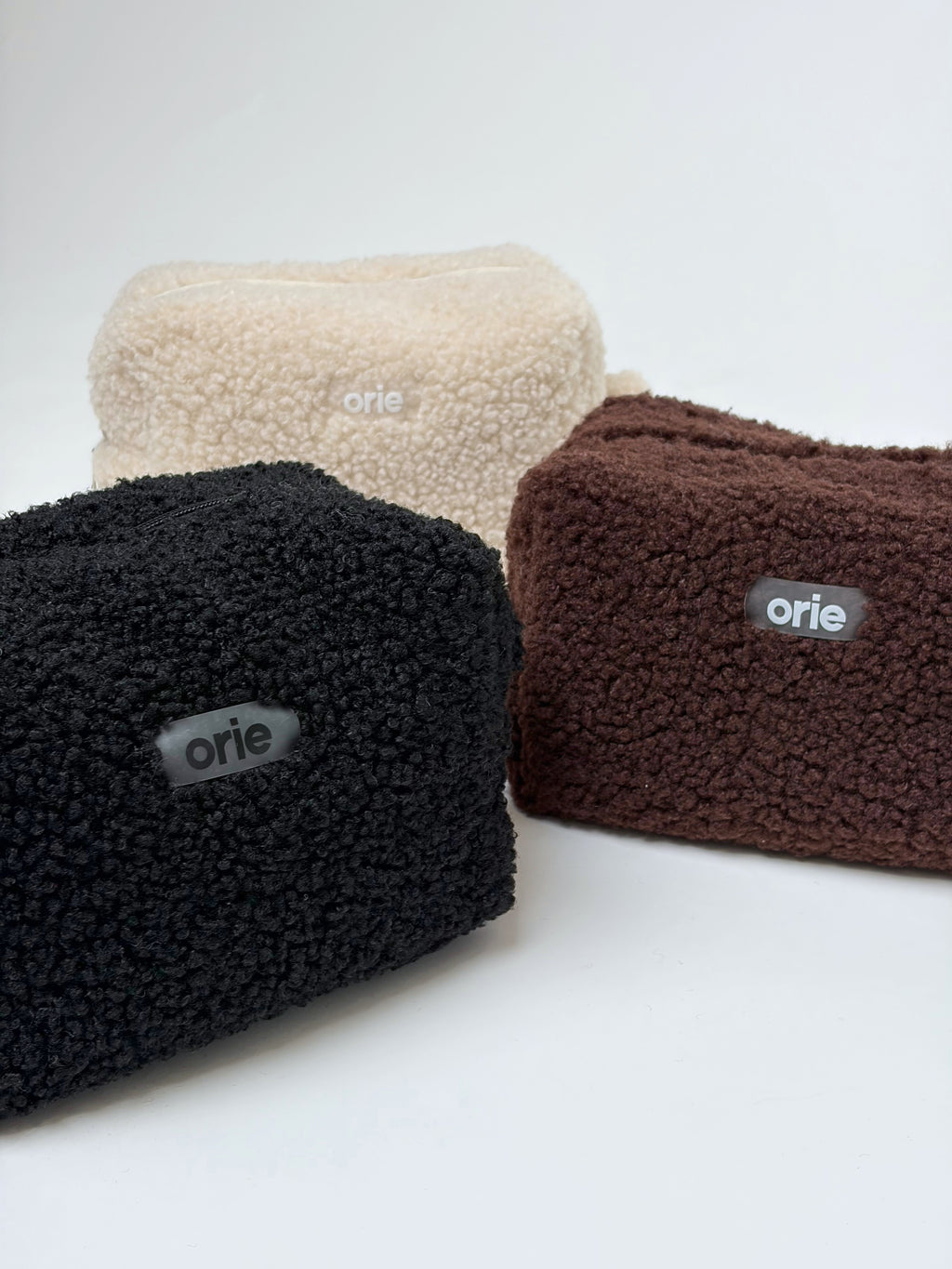 Four plush makeup bags in beige, black, and brown with 'orie' branding on a white background